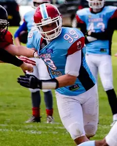 Adam Raine in a light blue Farnham Knights jersey and a red and white helmet, lunging forward during a game on a grass field.